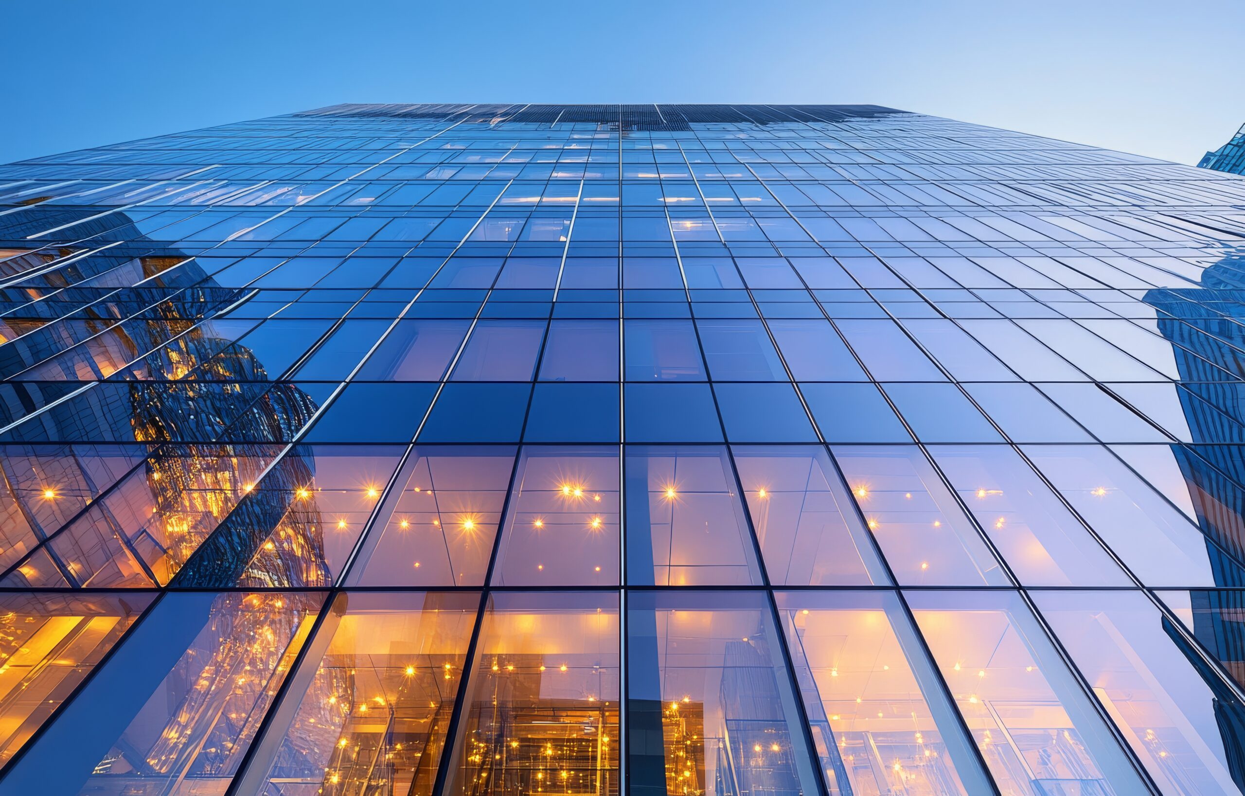 The sleek facade of a contemporary glass building showcases soft, warm lighting from the interior, creating a striking contrast against the deepening blue of the evening sky.